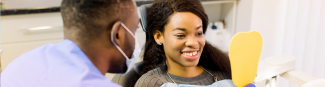 Une jeune femme noire regarde son sourire éclatant à l'aide d'un miroir jaune. Un hygiéniste dentaire est assis à côté d'elle.