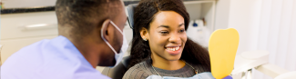 A young black female looking at her bright smile using a yellow mirror with a dental hygenist beside her