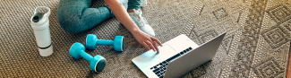 A close crop image of a woman sitting on the floor with her laptop open in front of her and weights and a water bottle beside her